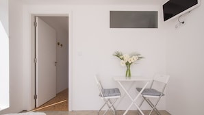 A small, modern room with a white door, a flat-screen TV mounted on the wall, a small table with a vase of flowers, and two foldable chairs. - CASASUPERTUBOS - APART T1 (Atouguia da Baleia)