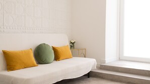 Ein weißes Sofa mit senfgelben und grünen Kissen, ein kleiner Beistelltisch mit Blumen in einer Vase und ein Fenster mit Blick ins Freie.