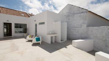 Un patio moderno al aire libre con paredes blancas, piso de concreto y techo de tejas. Se observan muebles blancos, incluyendo un banco y una mesita.