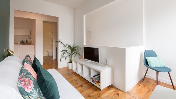 A modern living room with a flat-screen TV on a white entertainment unit, a blue chair, a potted plant, and a bed with floral pillows.