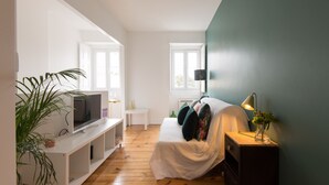 A bedroom with a bed, a TV on a white stand, a nightstand with a lamp, and a window.