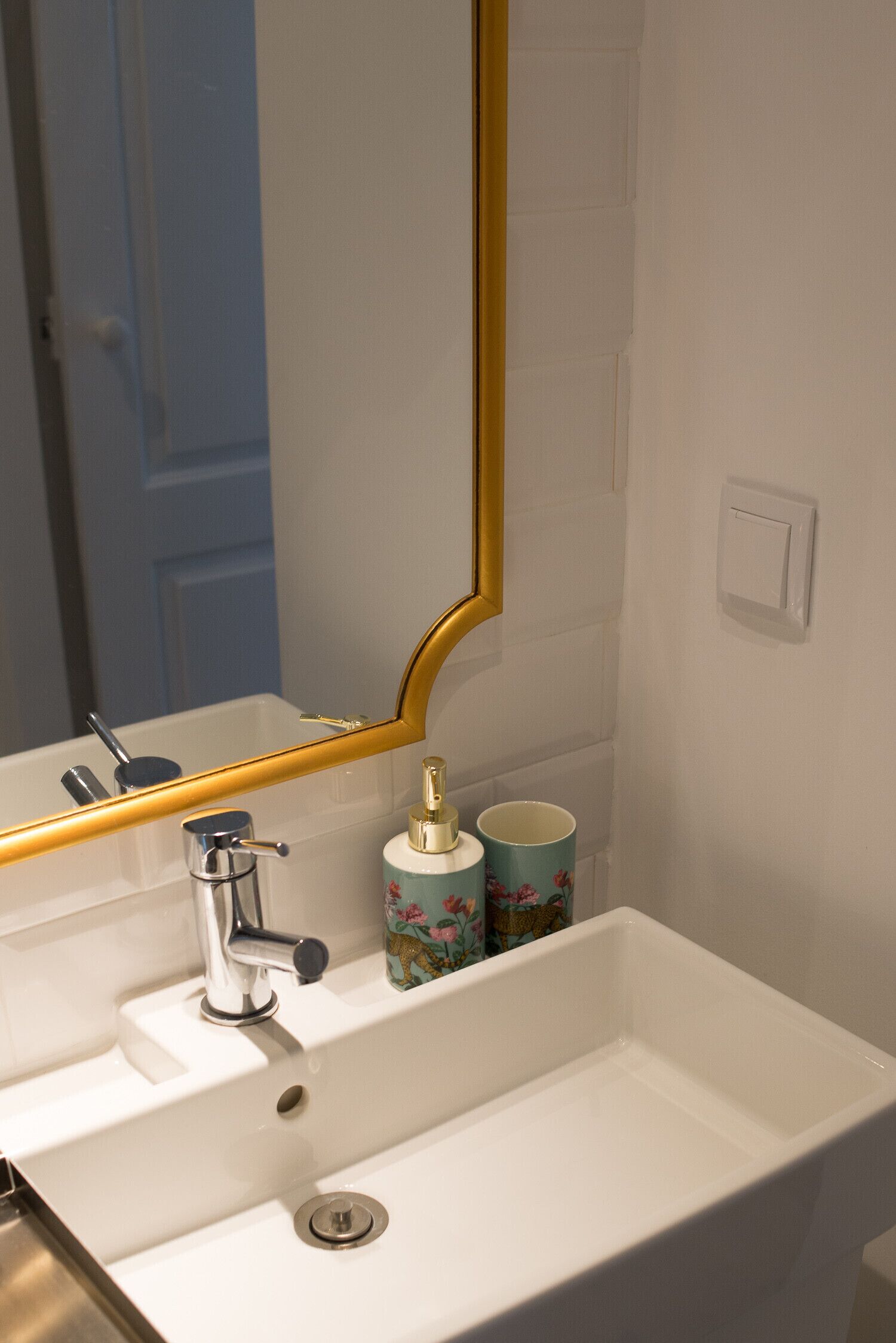 Un baño con un lavabo blanco, un espejo con marco dorado y dos tazas de estampado floral sobre el mostrador.