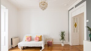 Un dormitorio con una cama blanca, suelo de madera, una araña, un radiador, una mesita con una lámpara y una planta en maceta.