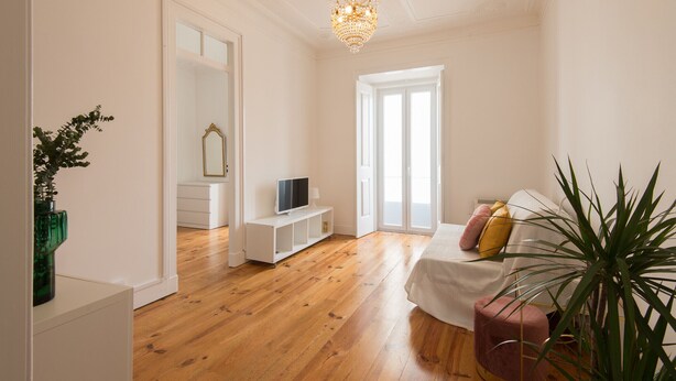 Una habitación amplia con suelo de madera, un sofá blanco, un televisor sobre un soporte blanco y un espejo grande.