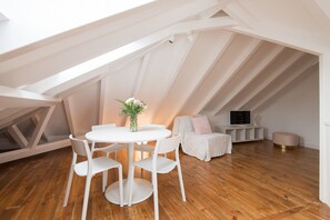 A high-ceilinged room with a wooden floor, a round table with chairs, a small sofa, and a television on a stand.