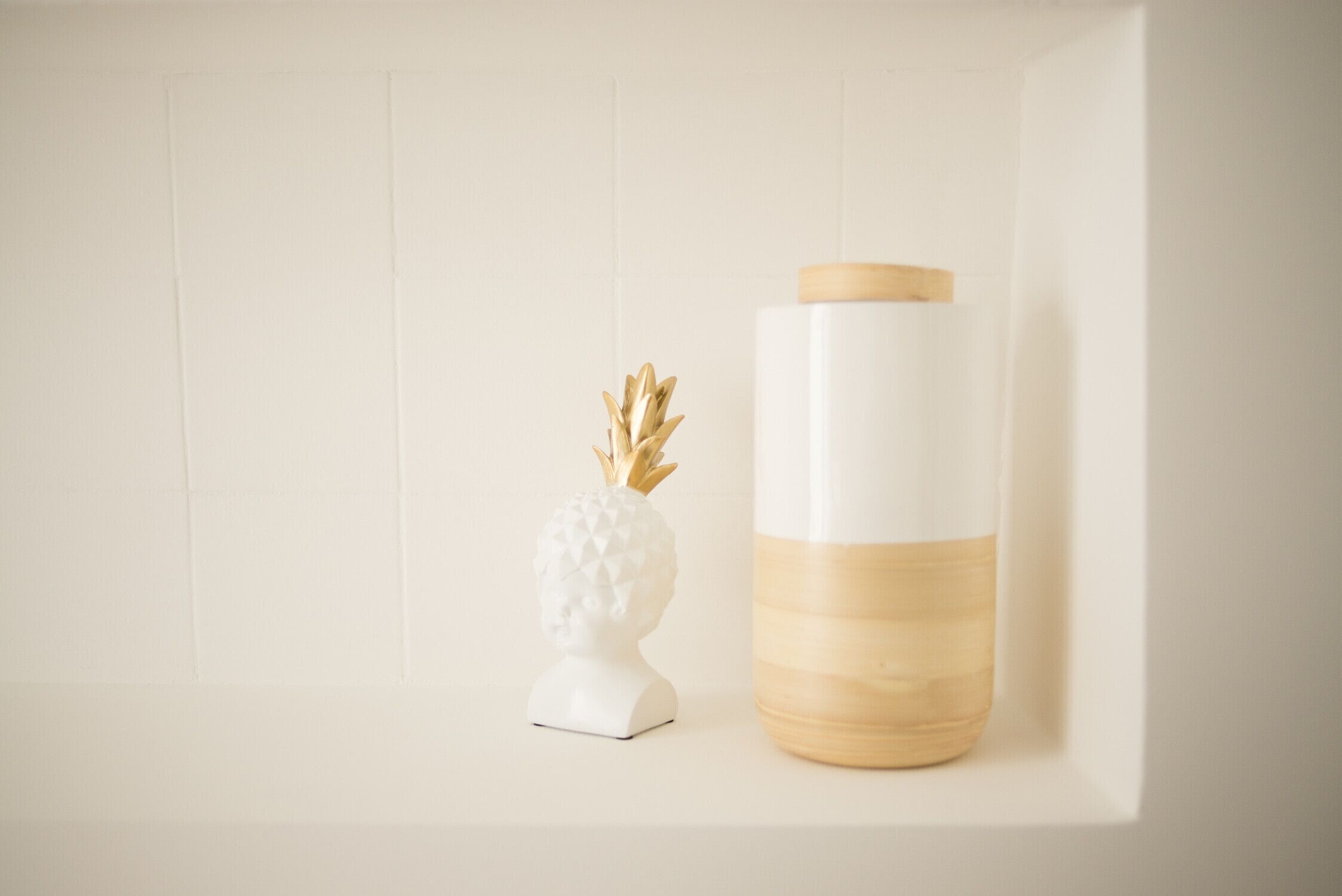 Eine weiße, ananasförmige Figur und ein weißer, aus Holz gefertigter Behälter auf einem Regal vor einer weißen Wand.