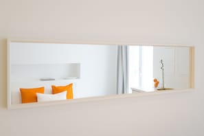 A hotel room with a large mirror, a bed with orange and white pillows, a small table with a vase, and a window with curtains.
