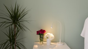Ett litet bord vid sängen med en vit lampa, en vas med röda och vita blommor och en hög, grön växt.