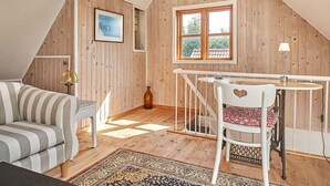A cozy room with a sloped ceiling, wooden walls, a striped sofa, a white chair with a heart design, a small table, and a window with a view of greenery. - 6 Person Holiday Home in Aakirkeby (Akirkeby)