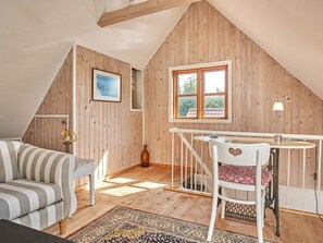 A cozy room with a sloped ceiling, wooden walls, a striped sofa, a white chair with a heart design, a small table, and a window with a view of greenery. - 6 Person Holiday Home in Aakirkeby (Akirkeby)