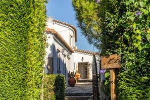 Un étroit sentier pavé de pierres menant à un bâtiment blanc surmonté d’un toit en tuiles, le tout entouré d’une végétation luxuriante.