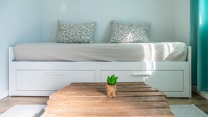 Une chambre à coucher avec un lit, deux oreillers, une table en bois avec une plante et une fenêtre avec des rideaux.