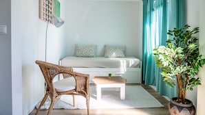 Une petite pièce bien éclairée, avec un lit, une chaise en osier, une petite table sur laquelle est posée une plante, et un arbre en pot.