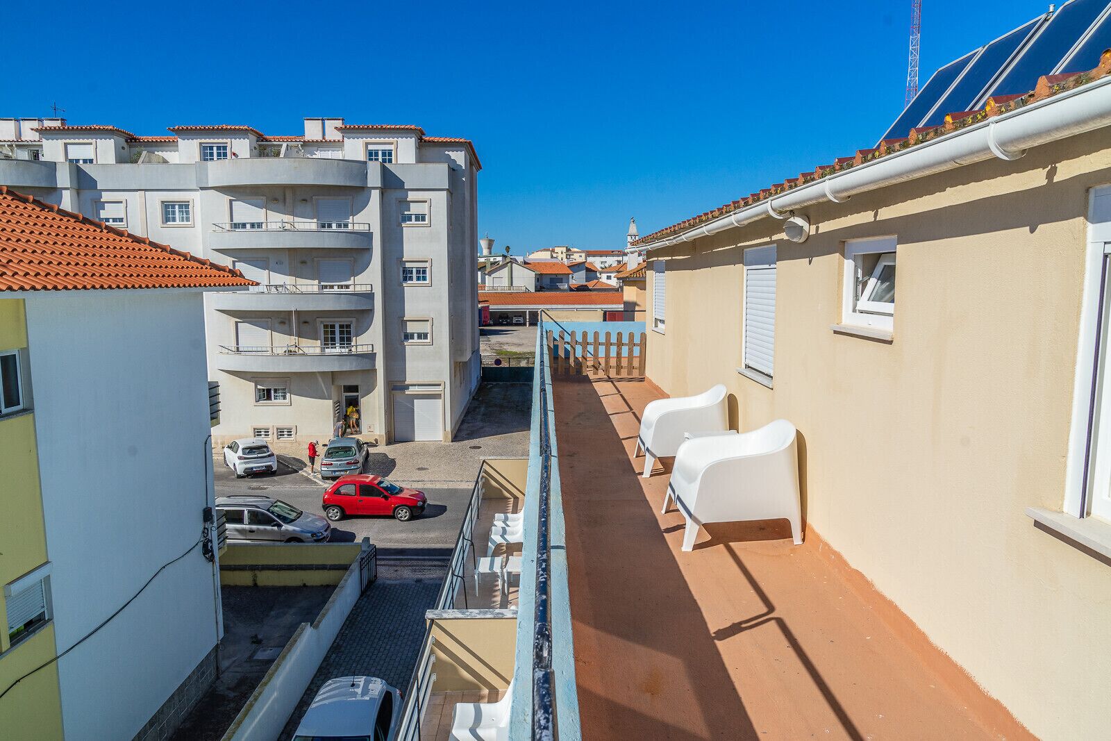 Un balcon étroit donnant sur une rue, des chaises en plastique blanches et une voiture rouge garée en contrebas.