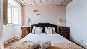 Un lit bien fait, avec du linge de lit blanc et deux serviettes enroulées, une tête de lit sombre et deux tables de chevet avec des lampes.