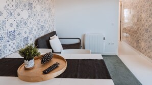 A modern room with patterned tile walls, a bed with a black headboard, a small table with a plant and remote control, and a radiator. - Encosta d'Óbidos T1 (Óbidos)