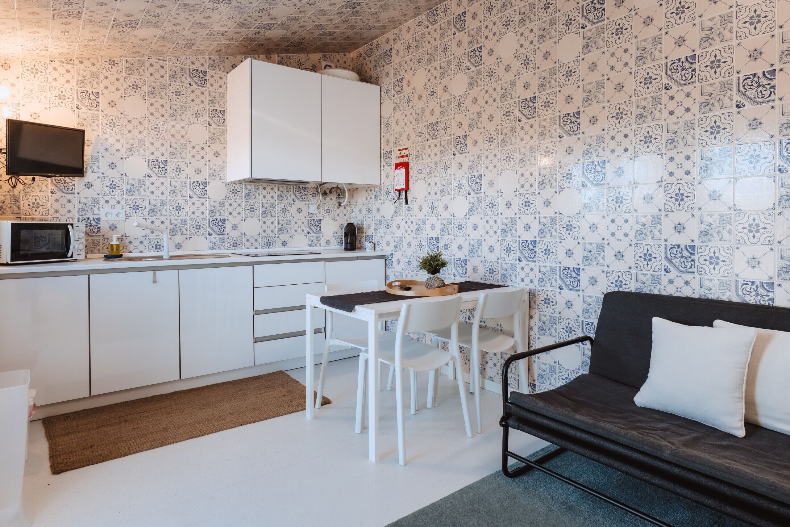 Een kleine keuken met witte kasten, een magnetron en een koffieapparaat. Er is een eettafel met stoelen en een donkere bank met witte kussens.