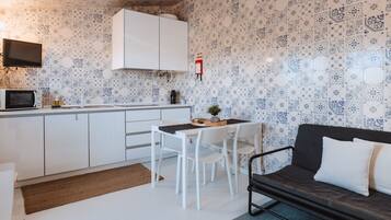 A compact kitchen with white cabinets, a microwave, and a coffee machine. There is a dining table with chairs and a dark sofa with white cushions.