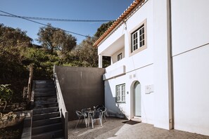 A white building with a tiled roof, a staircase leading to a door, and a small outdoor seating area with chairs and a table.