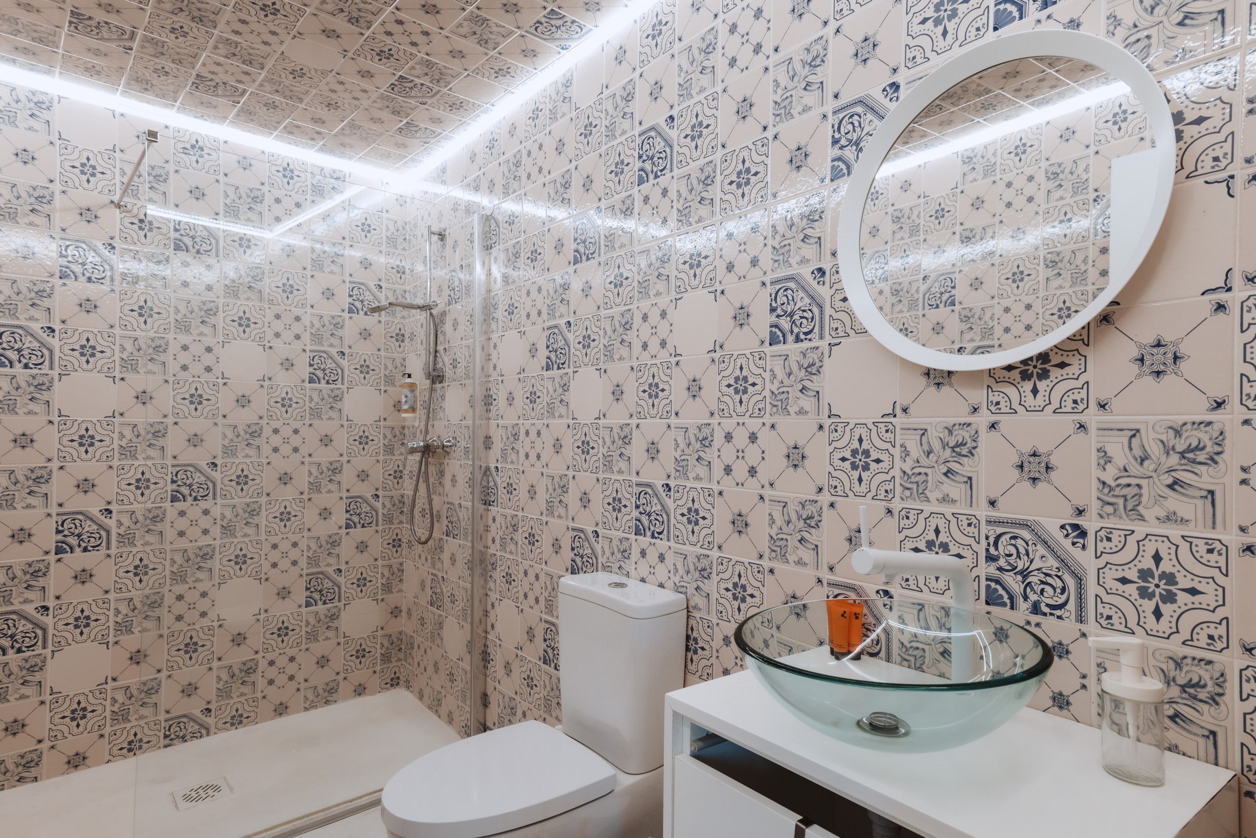 A bathroom with patterned tiles, a toilet, a glass sink with a mirror, and a shower area.