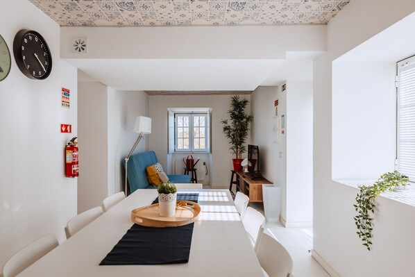A modern living room with a dining table, chairs, a blue sofa, a potted plant, and a window with blinds.