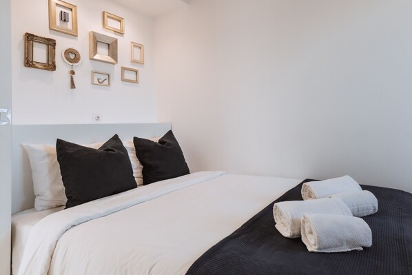 A neatly made bed with white and black bedding, a headboard, and a wall with various framed pictures.