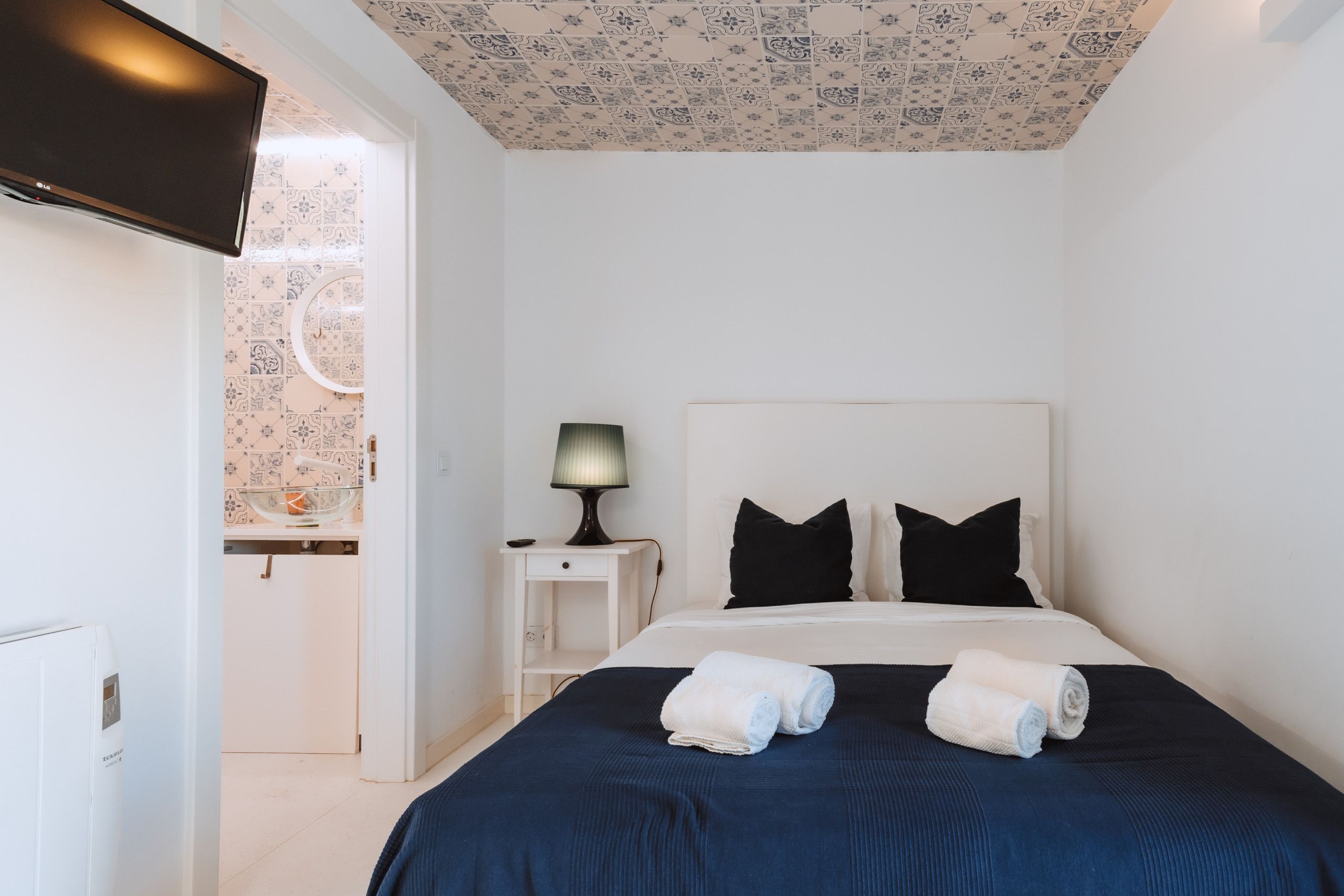 A hotel room with a bed, a nightstand with a lamp, a TV, and a bathroom visible through an open door.