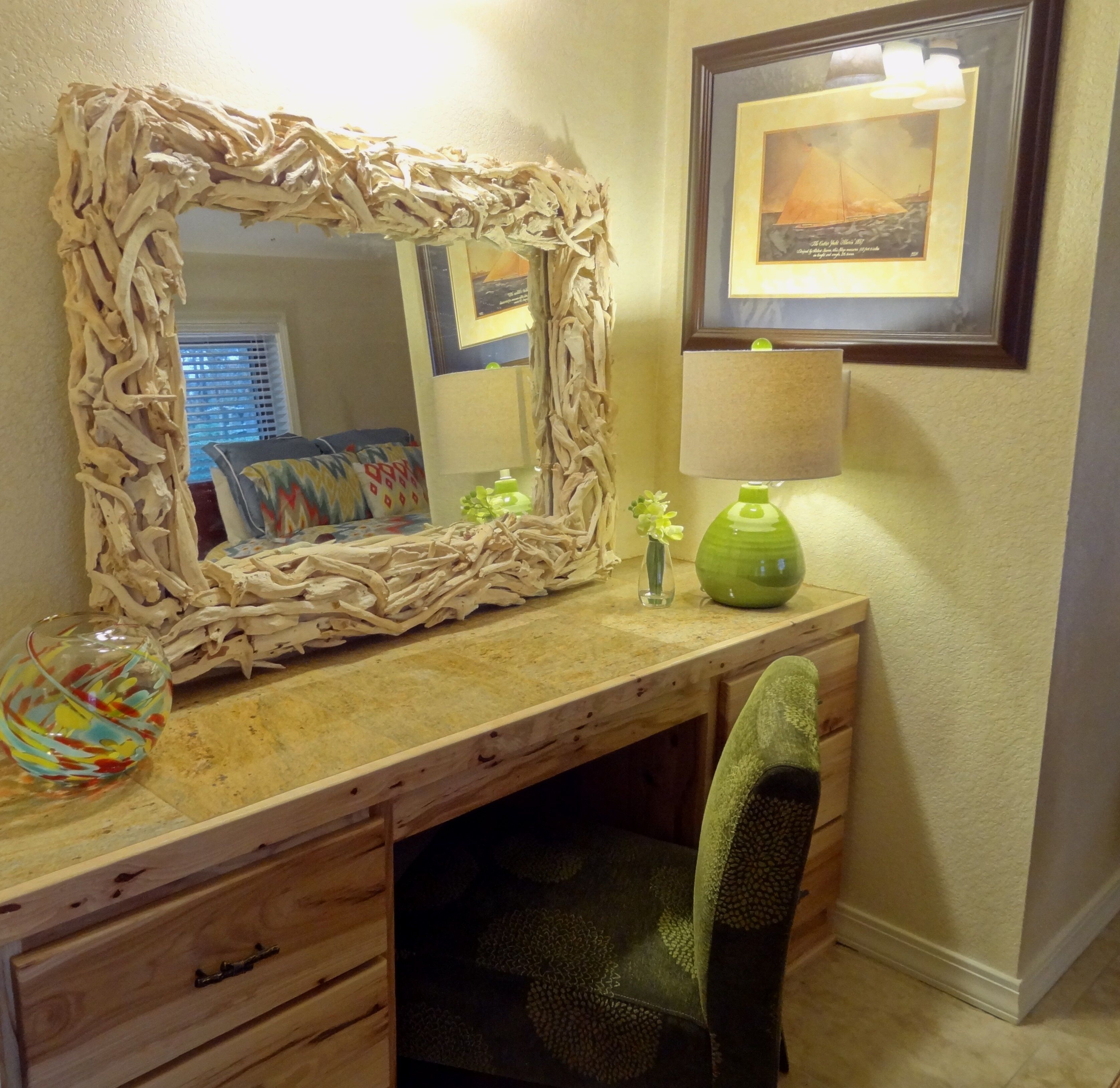 A bathroom with a wooden vanity, a mirror with a natural frame, a green vase, and a framed picture on the wall.