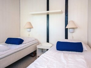 Habitació amb dos llits individuals, cada un amb un coixí blau i un matalàs blanc. Hi ha dues taules de nit, una a cada costat del llit, i dues làmpades penjades a la paret. Una estant blanca està adossada a la paret sobre els llits.