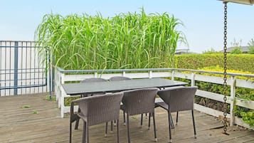 An outdoor patio with a table and chairs, surrounded by a white fence and tall green plants.