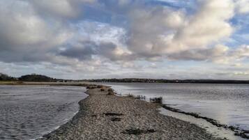 A coastal landscape with a pebbly beach, calm water, and a distant shoreline.