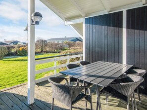 A covered outdoor area with a table and chairs, a white railing, and a view of a green landscape with houses in the background.