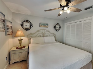 A bedroom with a bed, nightstand, lamp, ceiling fan, artwork, and closet.