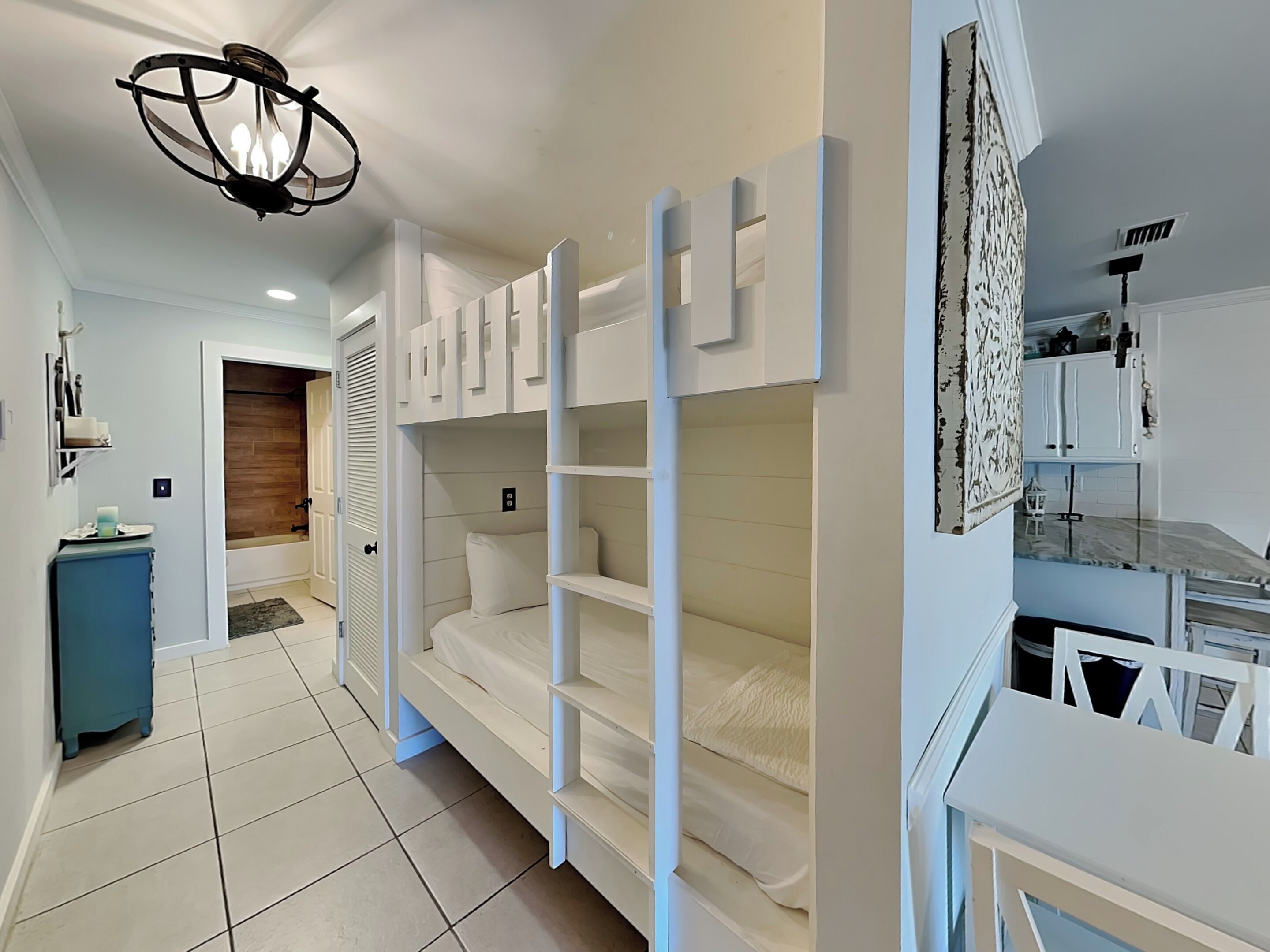 Une chambre avec des lits superposés, un bureau, une chaise, un luminaire au plafond et une porte donnant sur une autre pièce.