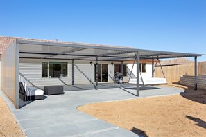A modern house with a covered patio area, a concrete driveway, and a wooden fence in the background. - Of Wine & Gods - Hot Tub, Bbq And Fire Pit! 3 Bedroom Home by RedAwning (Joshua Tree)