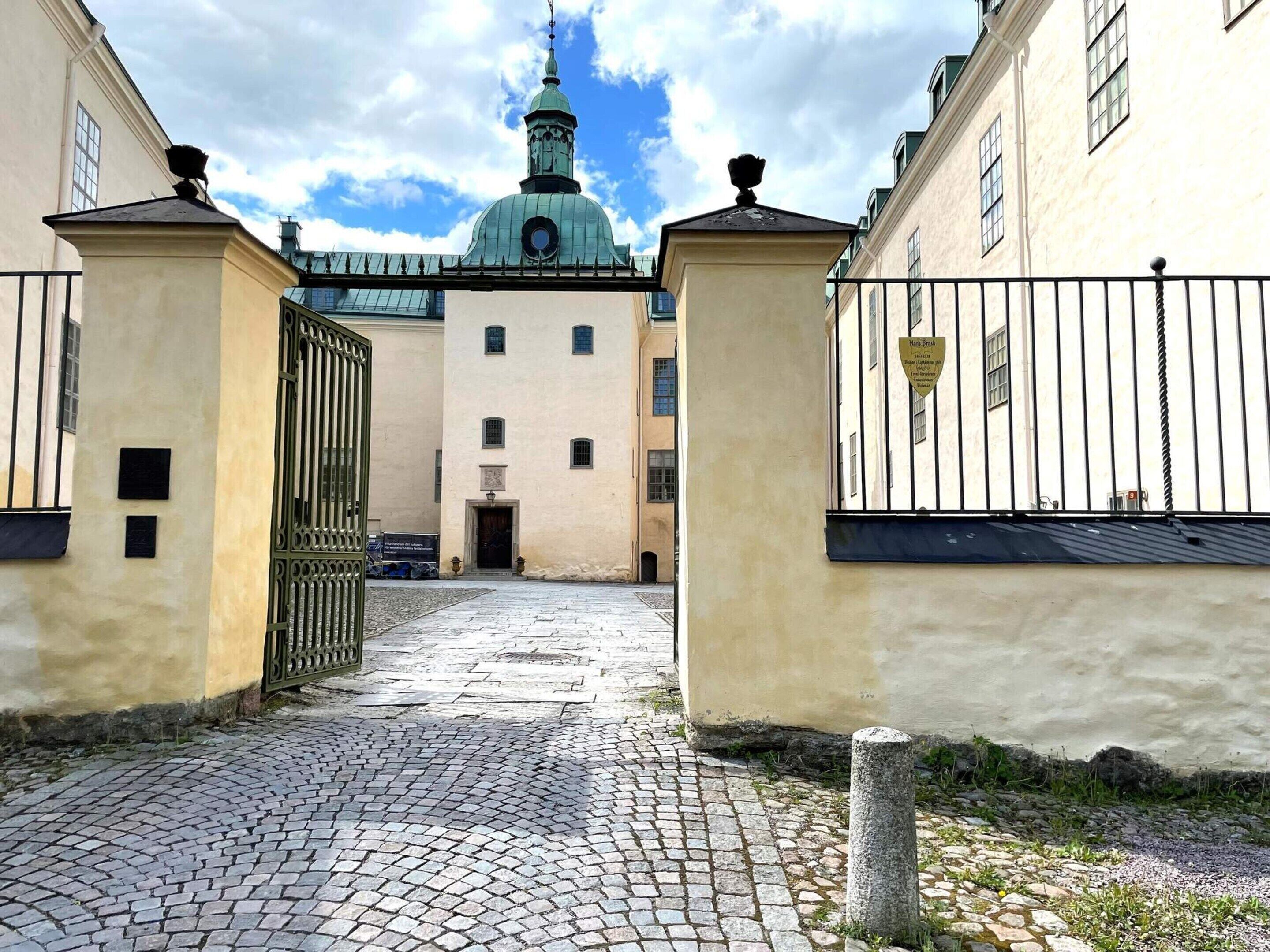 A cobblestone courtyard with a gate leading to a building with a dome and a clock tower.