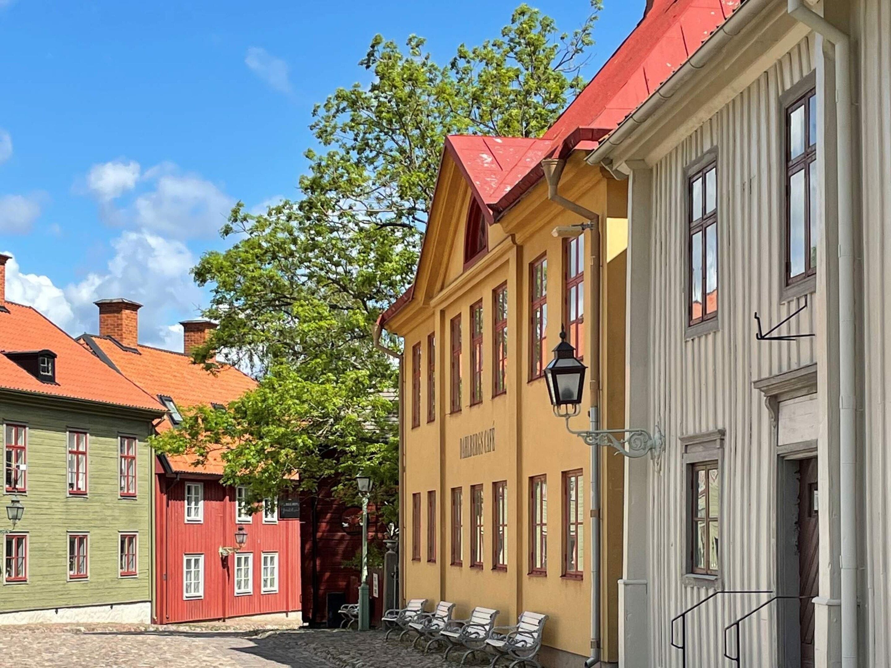 A cobblestone street with buildings featuring red and green facades, a yellow building with a red roof, and a row of empty chairs outside.