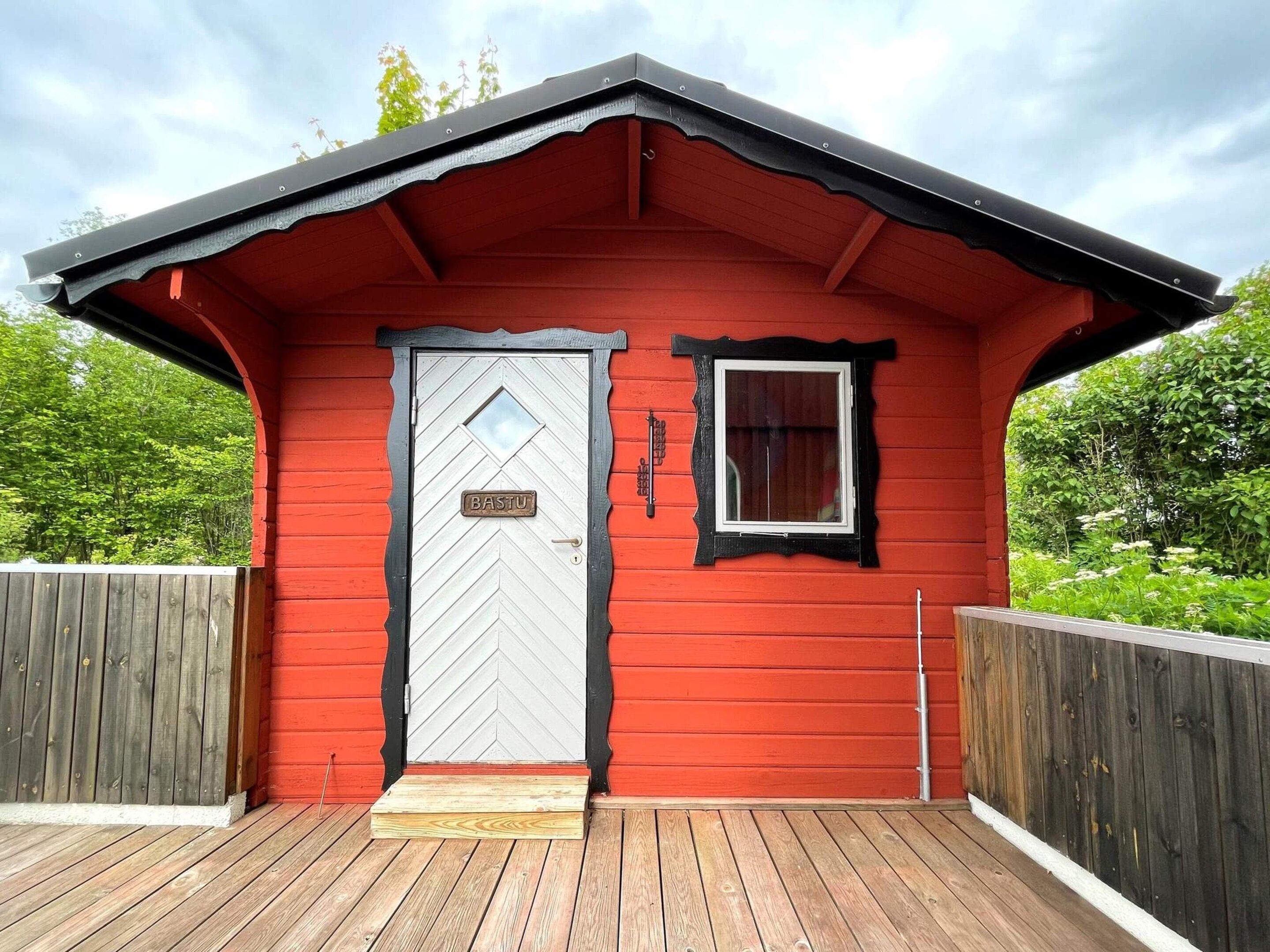 A red wooden cabin with a white door, a small window, and a wooden deck.