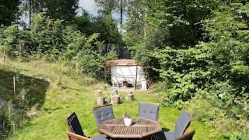 An outdoor seating area with a round wooden table and chairs, surrounded by lush greenery and a small structure with a tiled roof.