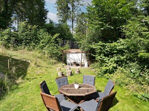 An outdoor seating area with a round wooden table and chairs, surrounded by lush greenery and a small structure with a tiled roof. - 6 Person Holiday Home in Melby (Melby)
