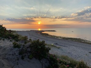 En strand med solnedgang, sandklitter og sparsom vegetation.