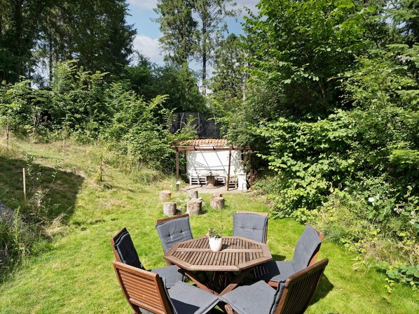 An outdoor seating area with a round wooden table and chairs, surrounded by lush greenery and a small structure in the background.