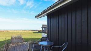 A wooden deck with a table and chairs, overlooking a grassy field and a building with a dark facade.