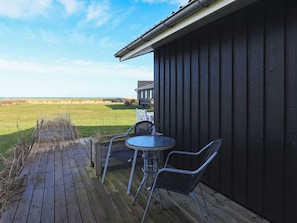 A wooden deck with a table and chairs, overlooking a grassy field and a building with a dark facade.