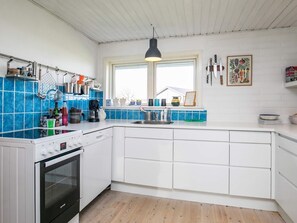 Et moderne køkken med hvide skabe, en indbygget ovn, et vindue med udsigt samt diverse køkkenredskaber og hårde hvidevarer.