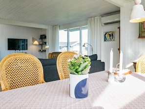 A dining area with a table set for two, a wicker chair, a sofa, and a television mounted on the wall.