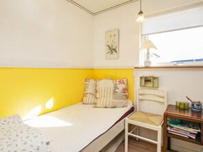 A single bed with a yellow wall, a bedside table with a lamp, and a small shelf with books and a cup.