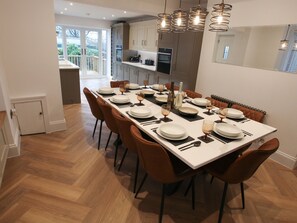 Un comedor con una mesa larga dispuesta para seis personas, rodeada de sillas marrones, en una cocina moderna con armarios blancos e iluminación colgante.