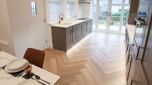 A modern kitchen with a central island, a dining area with a table set for four, and a sliding glass door leading to a balcony. - By the Tracks (Buxton)