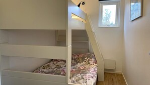 Un lit superposé avec une couvre-lit à motifs floraux, une fenêtre donnant sur les arbres et un luminaire fixé au mur.